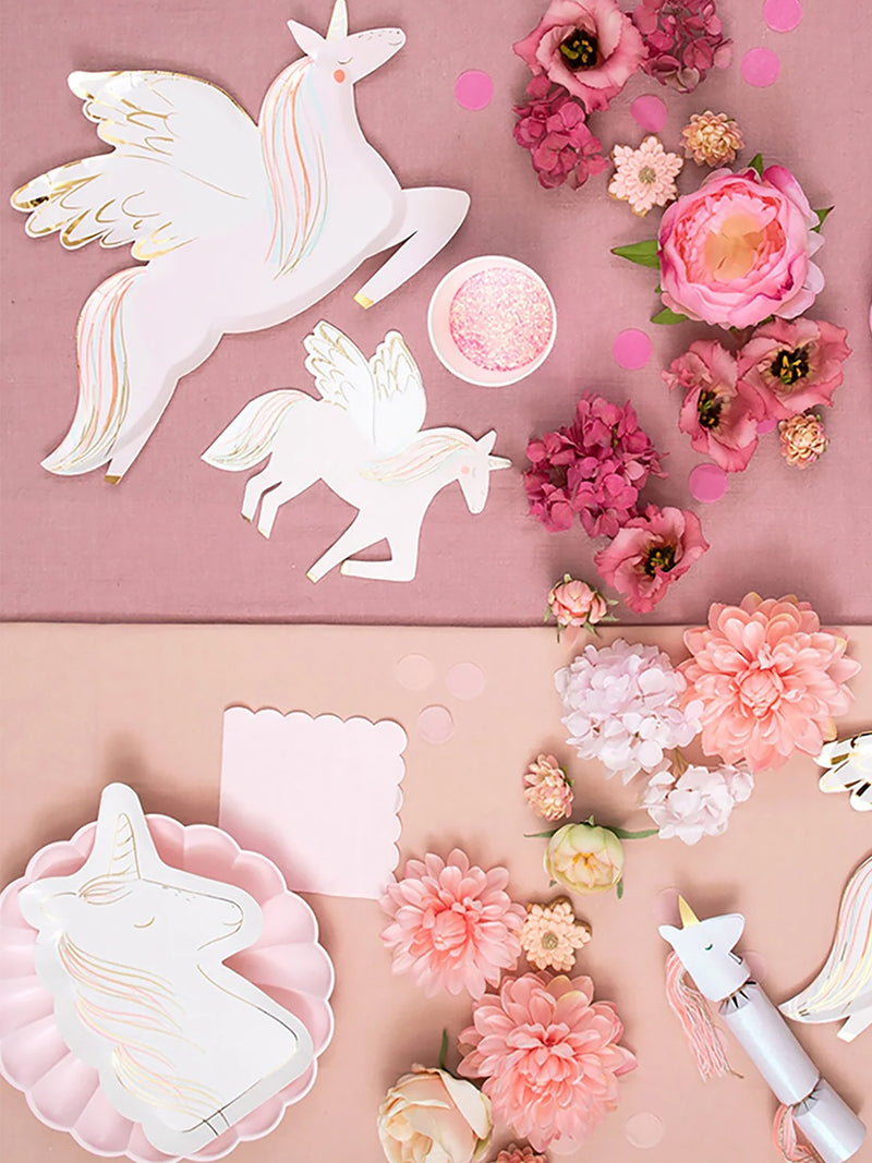 A styled flat-lay of unicorn-themed party decorations, including white and pink unicorn paper cutouts, matching unicorn disposable plates, pink flowers, scalloped pink cake napkins, and a small glitter bowl, arranged on a pink and peach background.