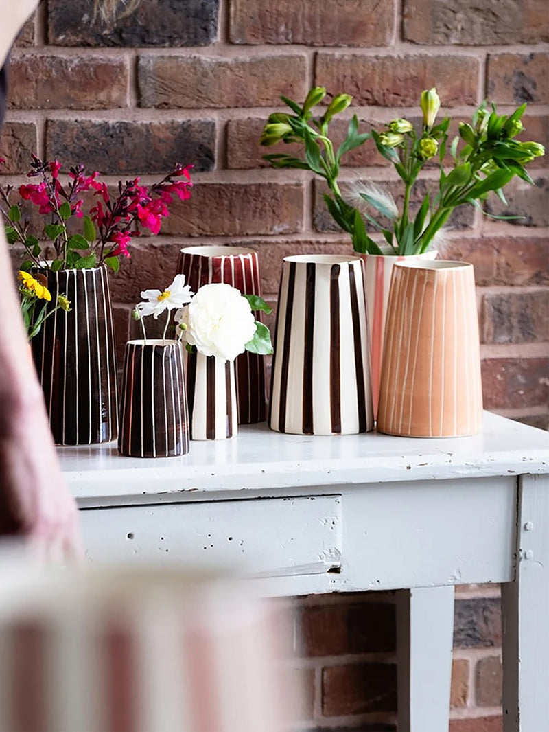A collection of six decorative ceramic vases and water jugs with distinct patterns (stripes, vertical lines, and ribbed textures) in black, white, brown, and peach, arranged on a white wooden table with flowers in front of a brick wall.