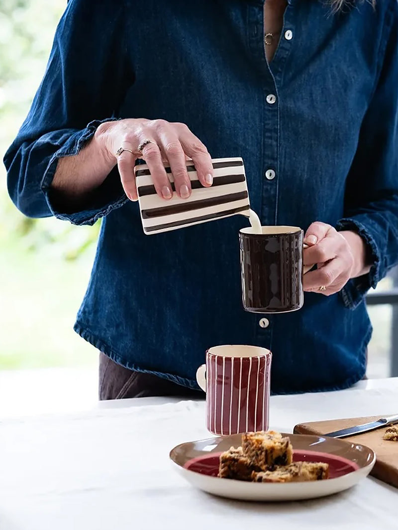 A person in a dark blue denim shirt pours milk from a striped cream and brown pitcher into a dark brown ceramic mug. Another striped mug and a plate with baked goods sit on a white table in the foreground.