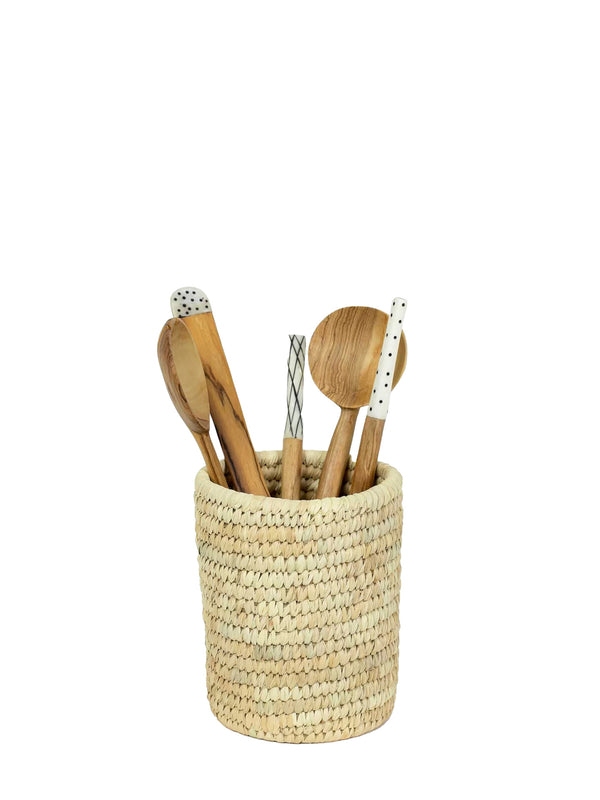 A woven natural fiber cylindrical holder filled with wooden kitchen utensils, including spoons and a whisk, on a plain white background.