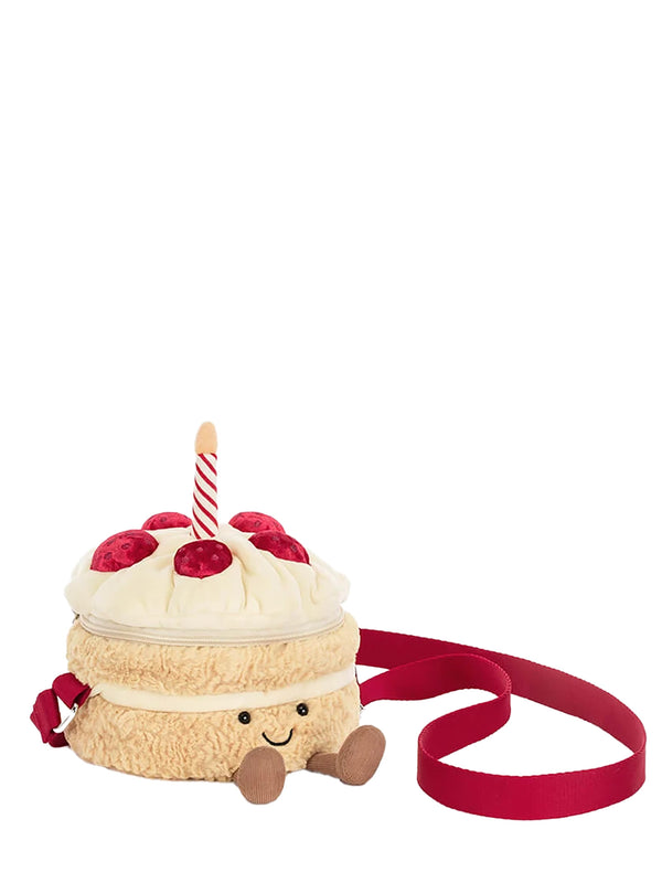 A plush cake-shaped shoulder bag with a cream-colored frosting top, red cherries, a single lit candle, and a red strap, shown on a plain white background.