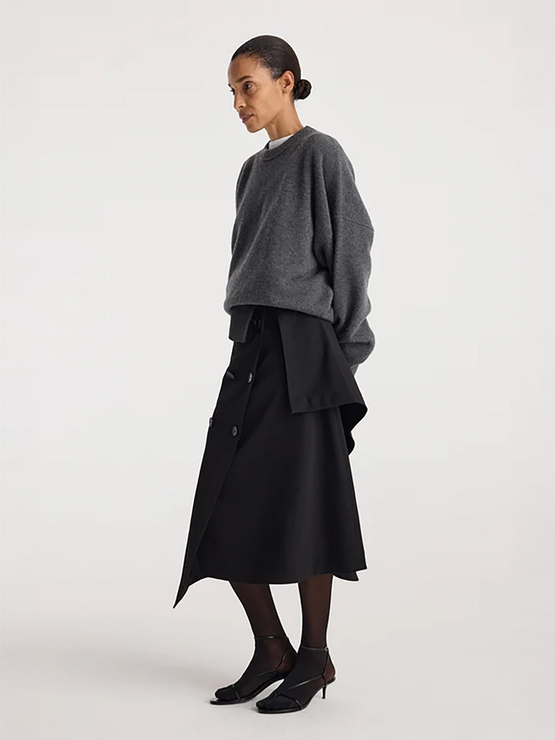 A model is shown in profile wearing a dark gray oversized knit sweater and a black asymmetrical midi skirt with buttons, paired with black tights and black ankle boots, against a plain light gray studio background.