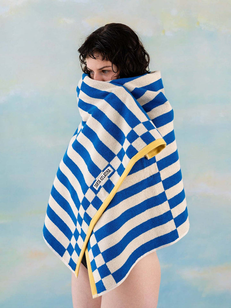 A person wrapped in a striped beach towel with blue and white stripes and a yellow trim, shown against a soft blue background. The towel has a visible 'Tarta Gelatina' label.