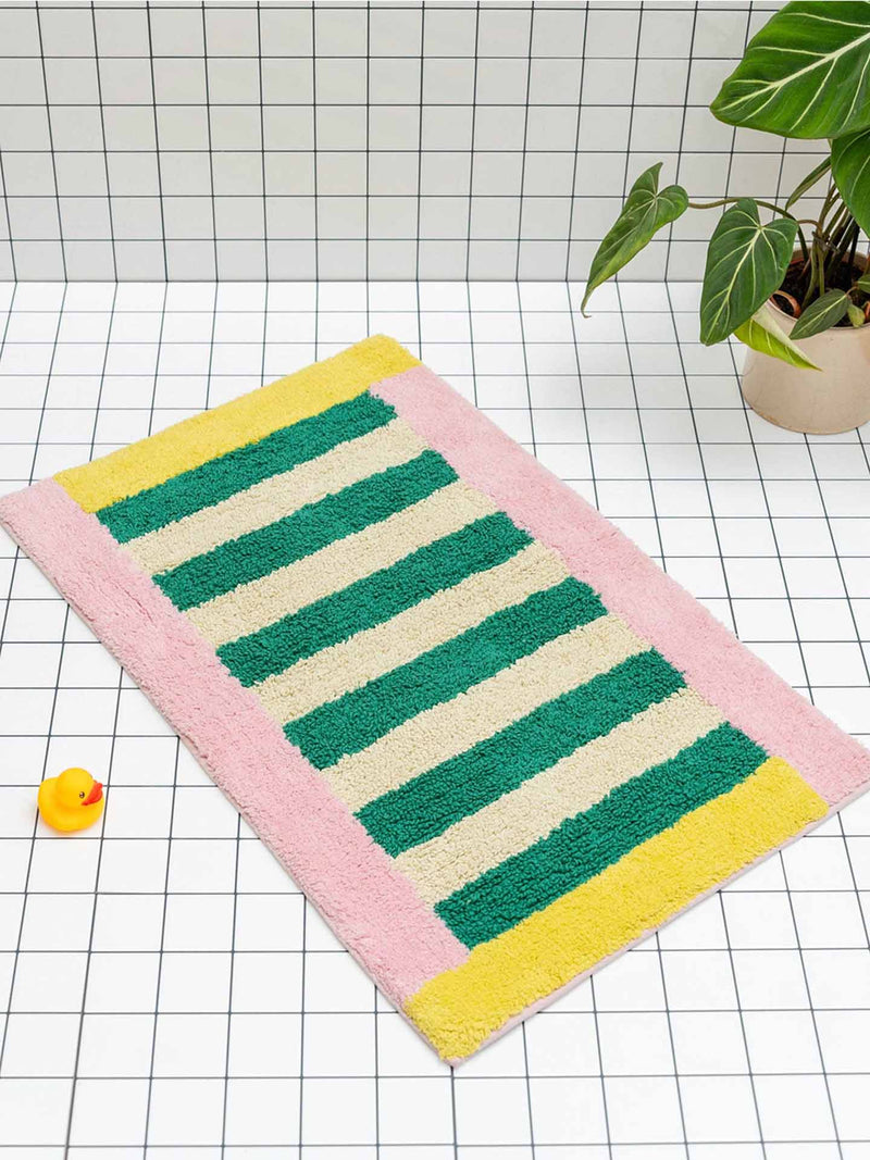 A rectangular bath mat with green, cream, and yellow stripes and a pink border, styled on a white tiled floor next to a small yellow rubber duck and a potted plant.