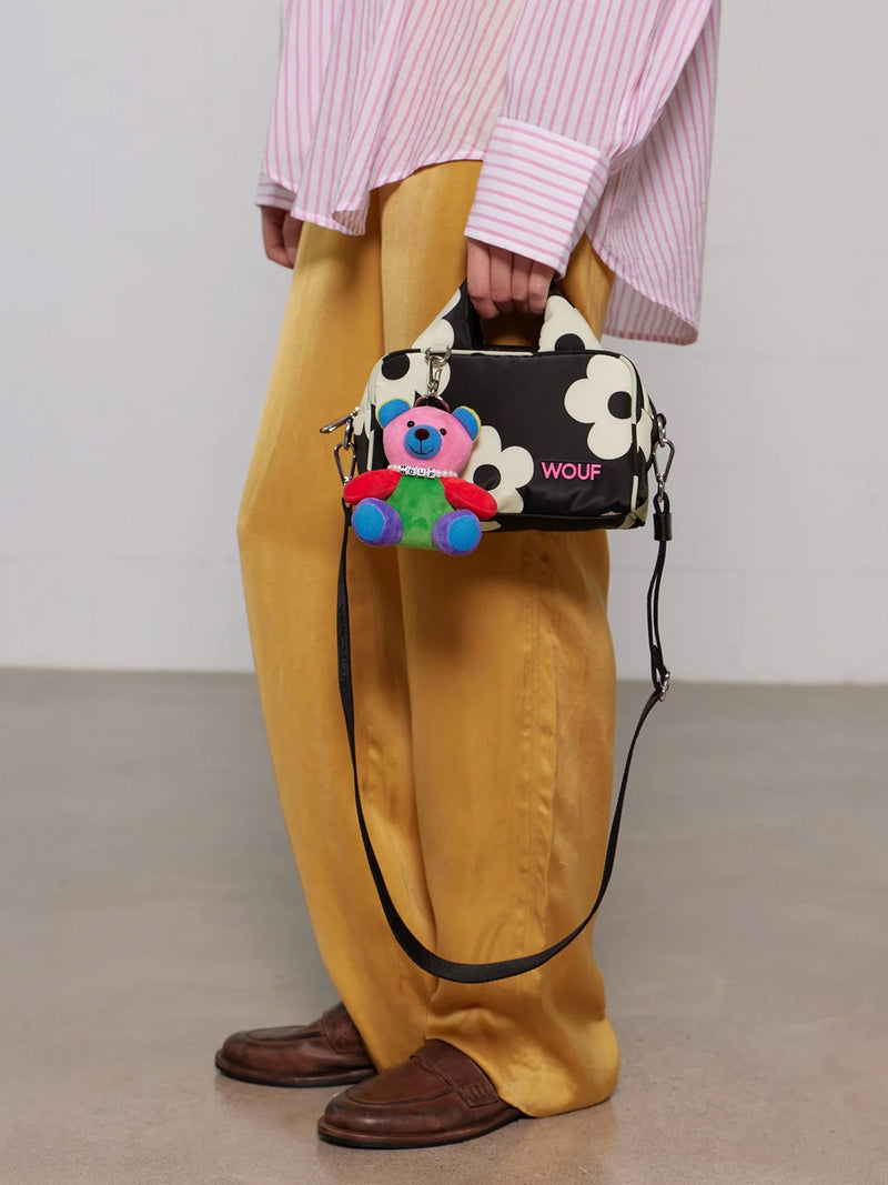 Person holding a black handbag with floral design and colorful bear toy, wearing mustard pants and pink shirt.