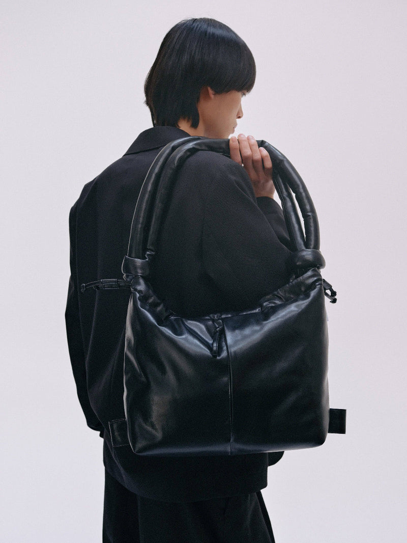 A male model holding a large black leather bag against a plain background