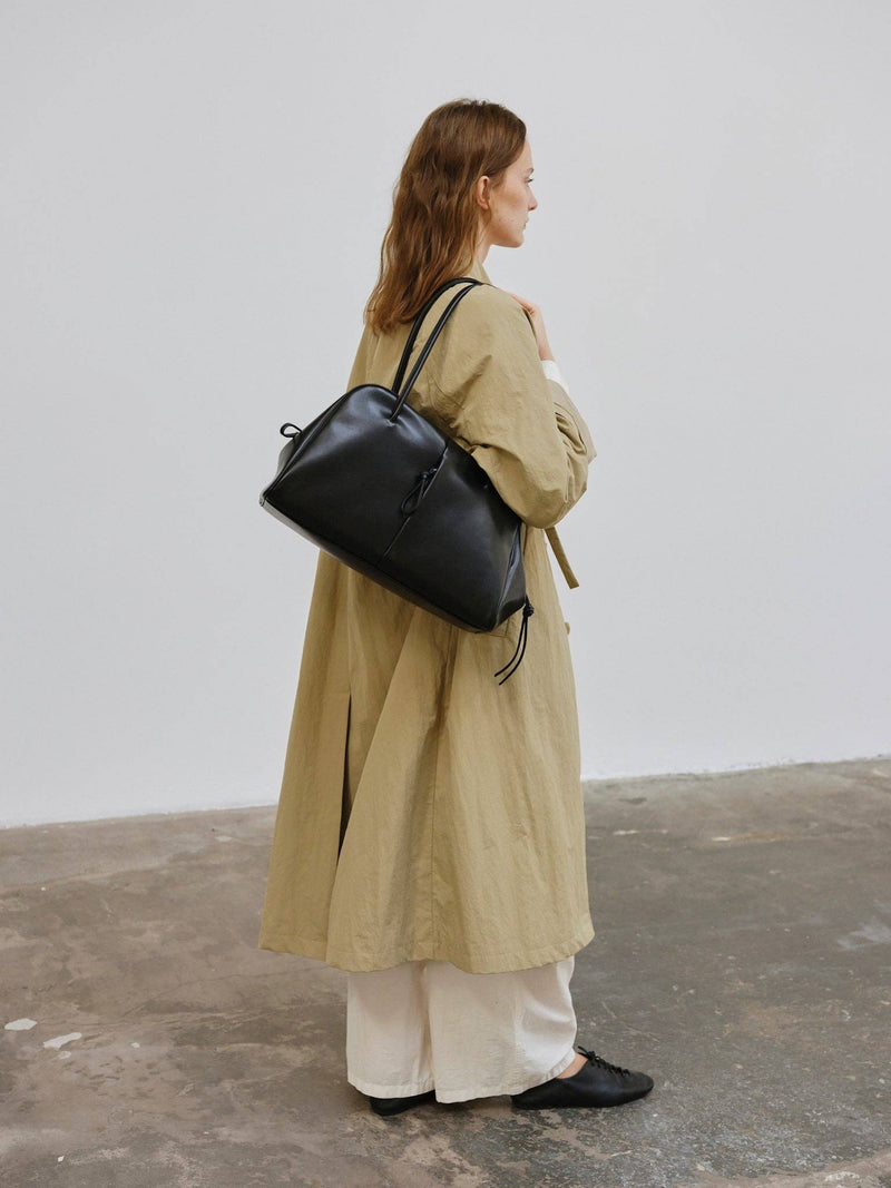 Person wearing a beige coat and holding a large black bag on her shoulder standing on a concrete floor, with a plain light grey wall behind