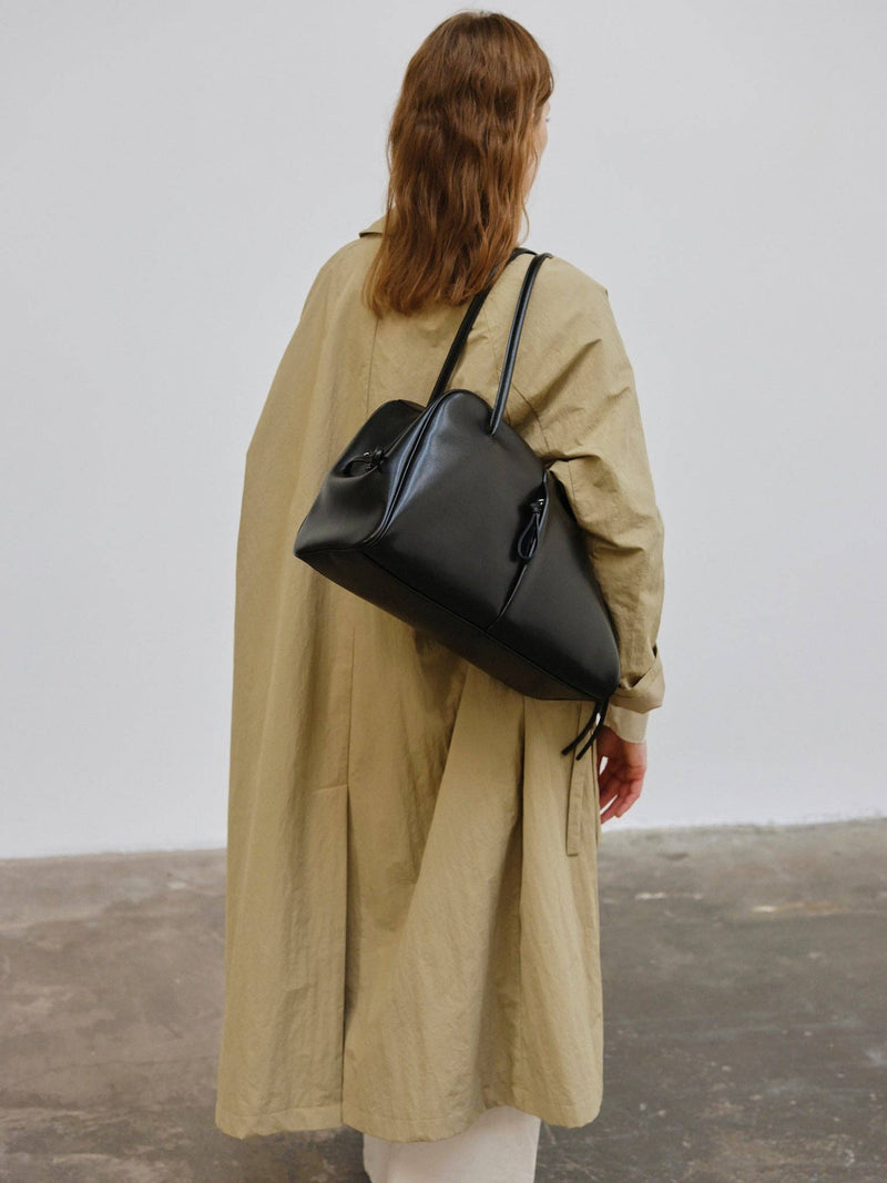 Person wearing a beige trenchcoat and carrying a medium-large black bag against a plain background