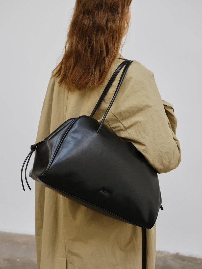 Person wearing a black shoulder bag over her shoulder with a beige coat and long red hair against a plain background