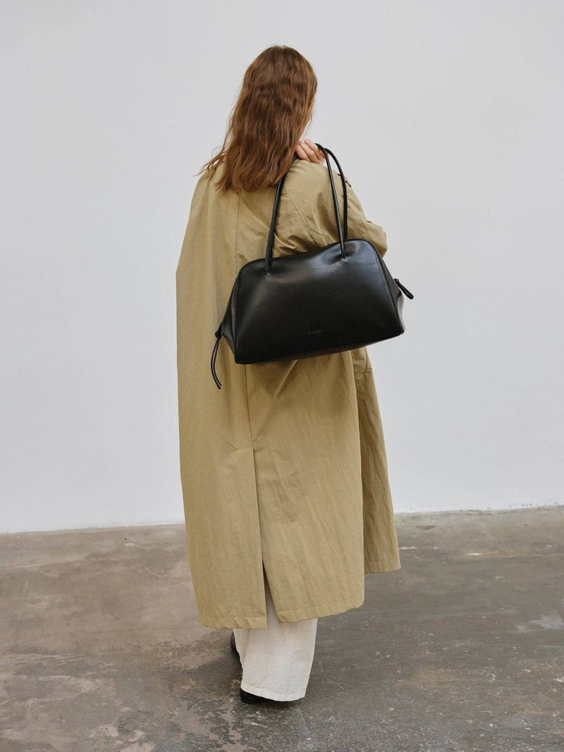 Person wearing a long beige coat and carrying a black bag against a plain background