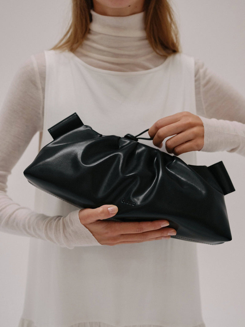 Person holding a black clutch bag and tightening a small string loop on the bag's top against a neutral background