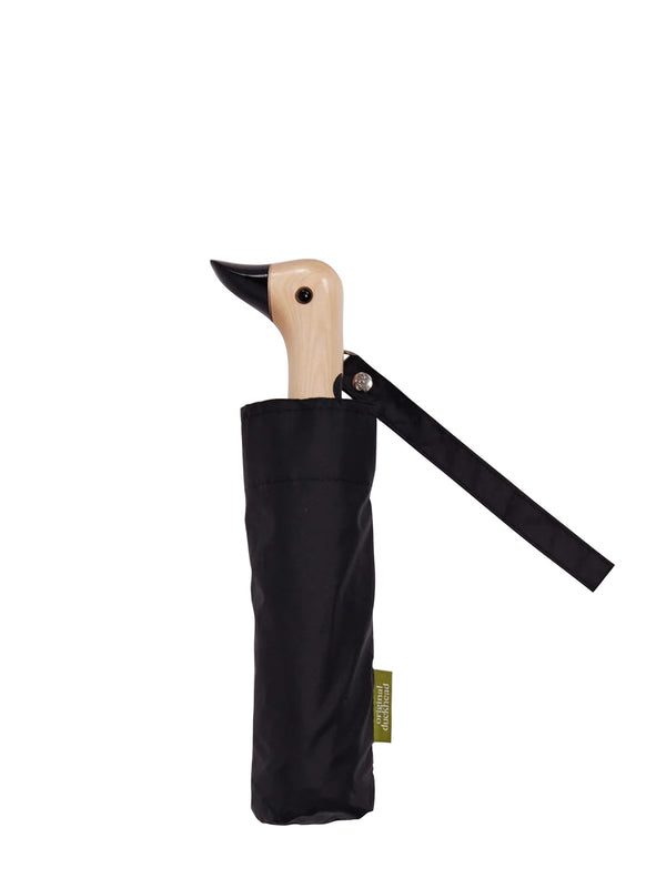 A folded black umbrella with a light natural wood handle in shape of a duck or bird, with round beady eye and black painted beak in a cylindrical black sleeve with a small green brand label 'Original Duckhead' and black hanging loop from the canopy material, shown on a plain white background.
