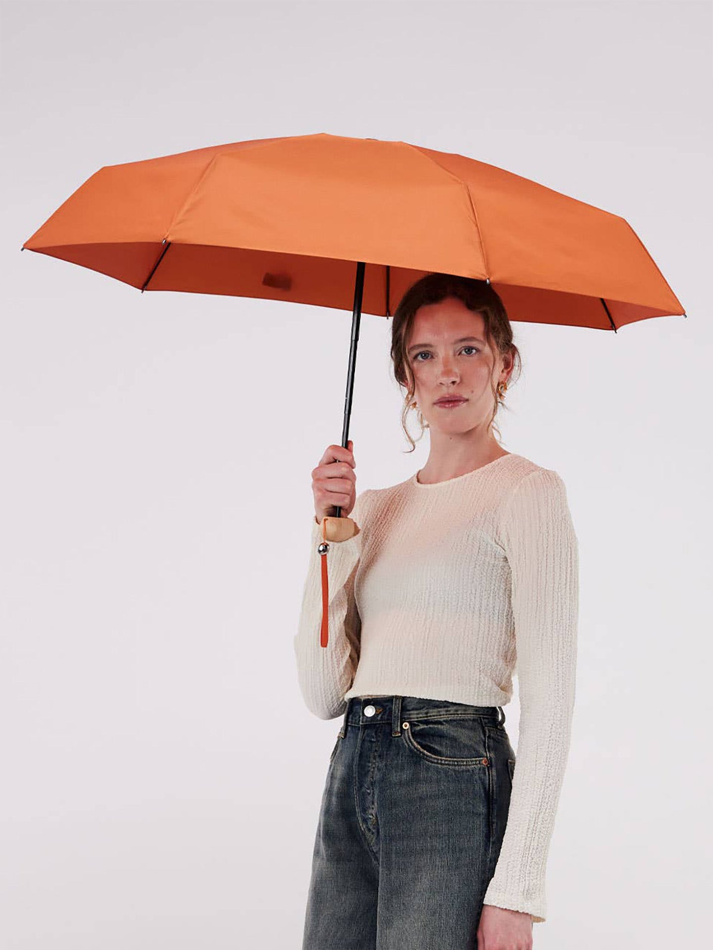 A woman holding a bright orange umbrella, wearing a cream ribbed long-sleeve top and dark gray jeans, against a plain light gray studio background.