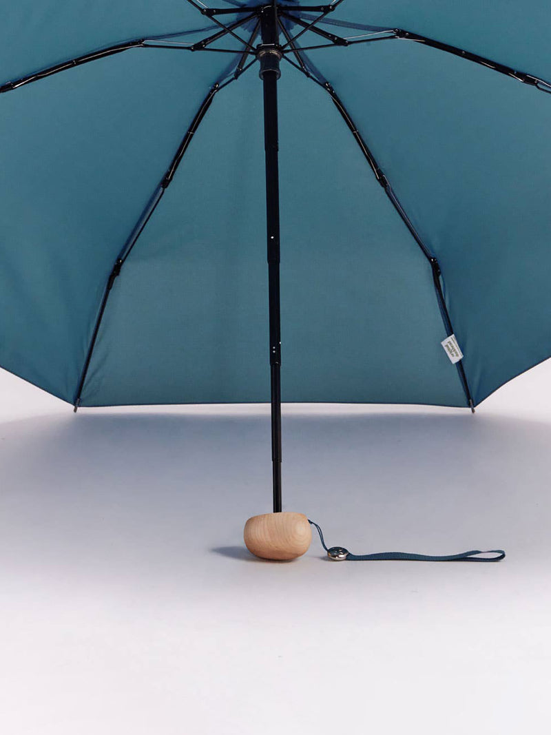 A teal blue folding umbrella with a black frame and a small oval wooden base, shown open against a clean white background.