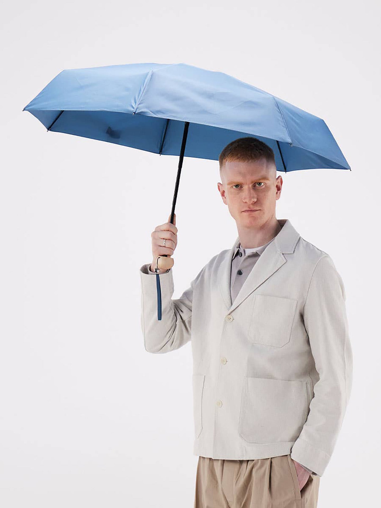 A man in a light beige blazer and khaki trousers holds up a light blue umbrella against a plain white studio background.