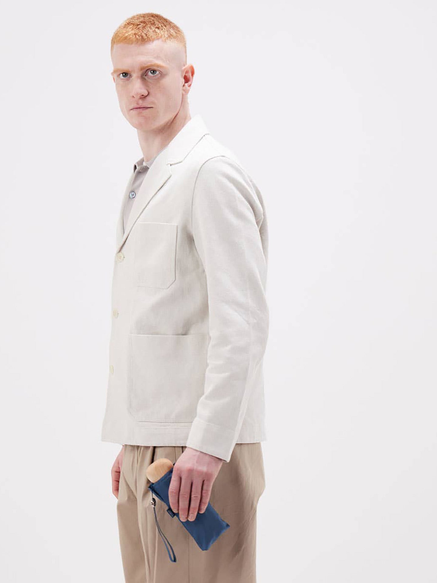 A man with red hair is shown in profile wearing a smart light beige blazer over a collared shirt and beige trousers, holding a dusty blue mini umbrella slipped in a matching blue pouch photographed against a plain white studio background.
