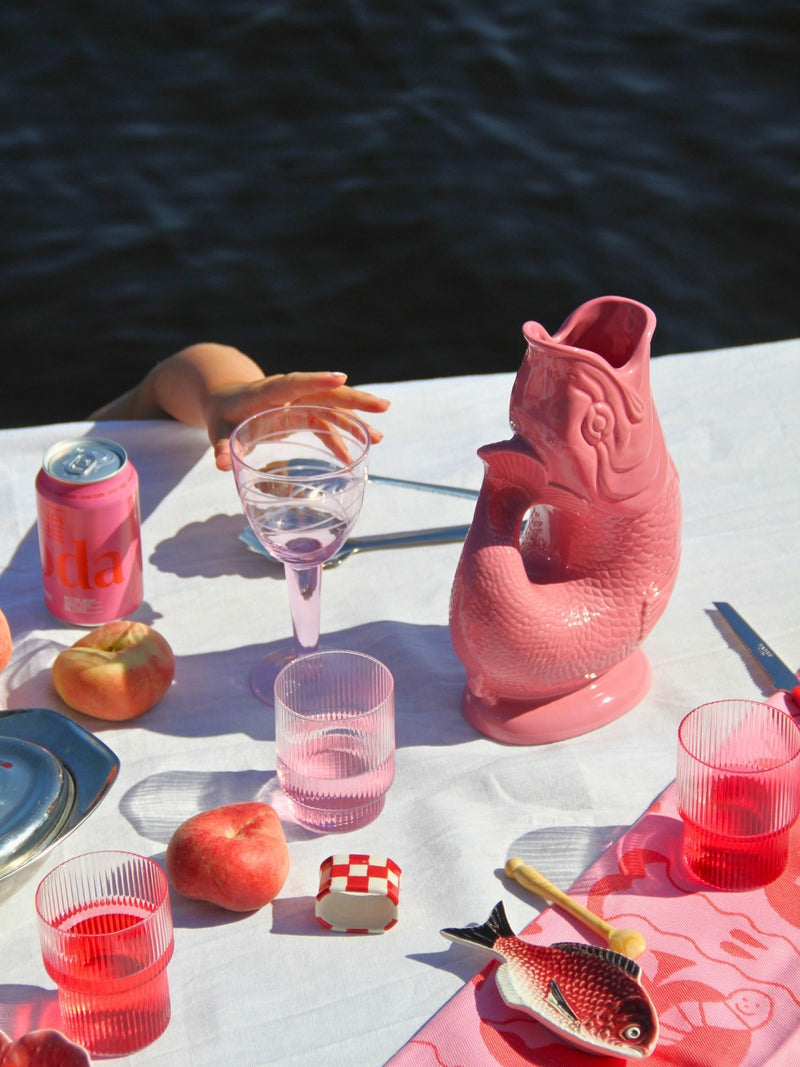A pink fish-shaped pitcher sits on a white tablecloth outdoors near water, surrounded by matching pink drinking glasses, a pink can, peaches, and a hand resting near the pitcher.