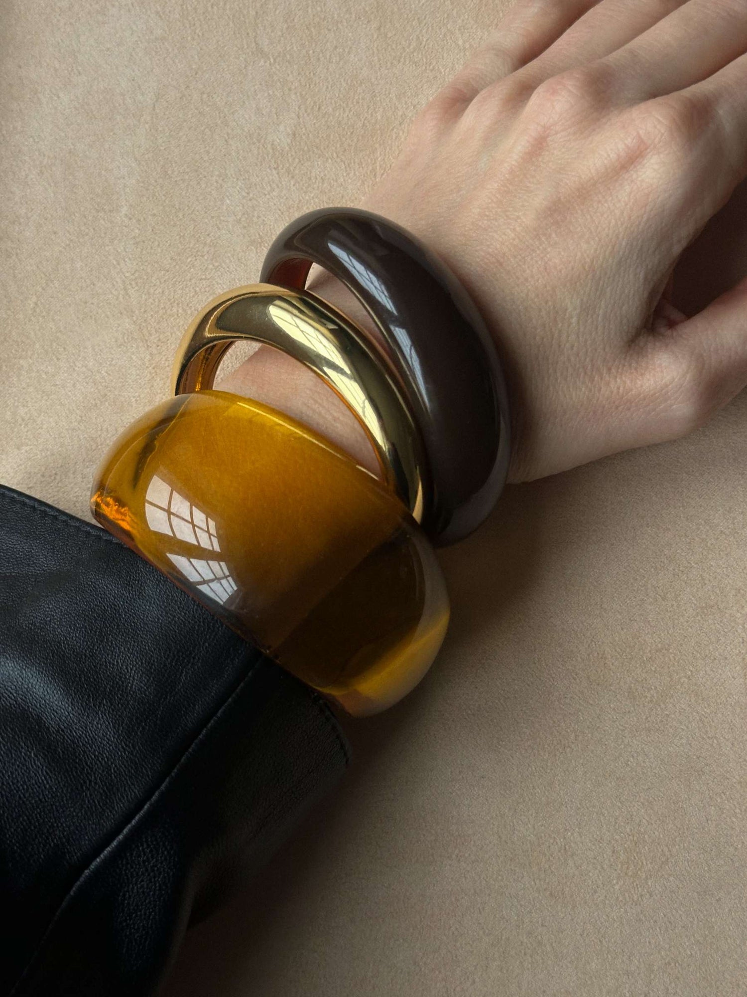 Close-up of a wrist wearing one wide resin bangle in see-through amber brown and two smaller bangles, gold and dark brown or black, resting on a beige background