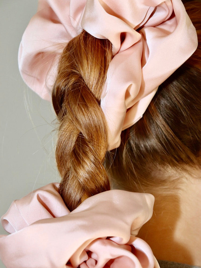 Back of the head photograph of a person in the sun hair lift up and gathered on a high ponytail by a big light pink shiny satin scrunchie and then braided and tied together by another big light pink shiny satin scrunchie.