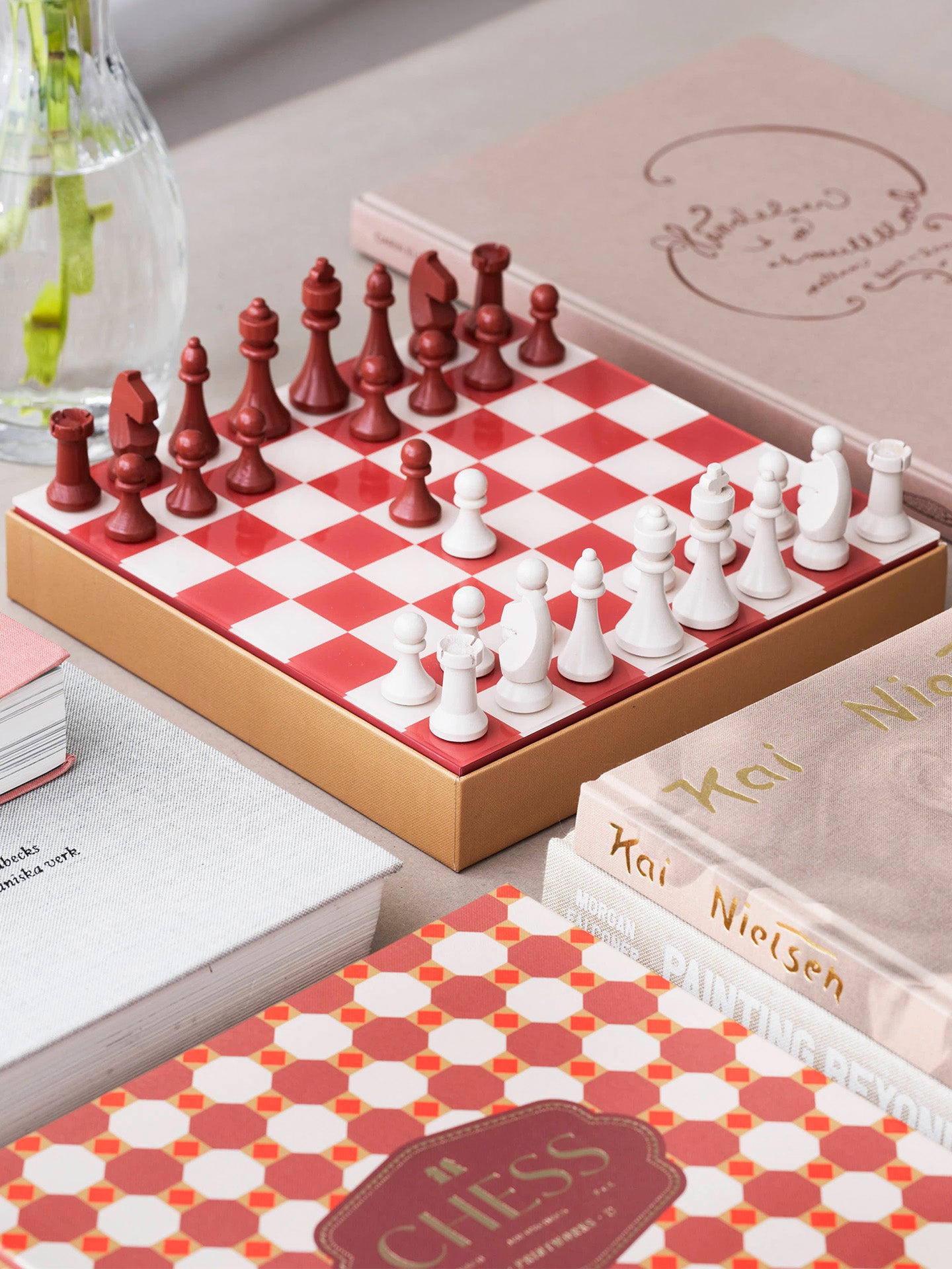 A boxed chess set with a glossy red-and-white checkered board and classical shaped white and dark red chess pieces, styled on a tabletop with the box cover, beautiful coffee table books and a glass flower vase in the background.