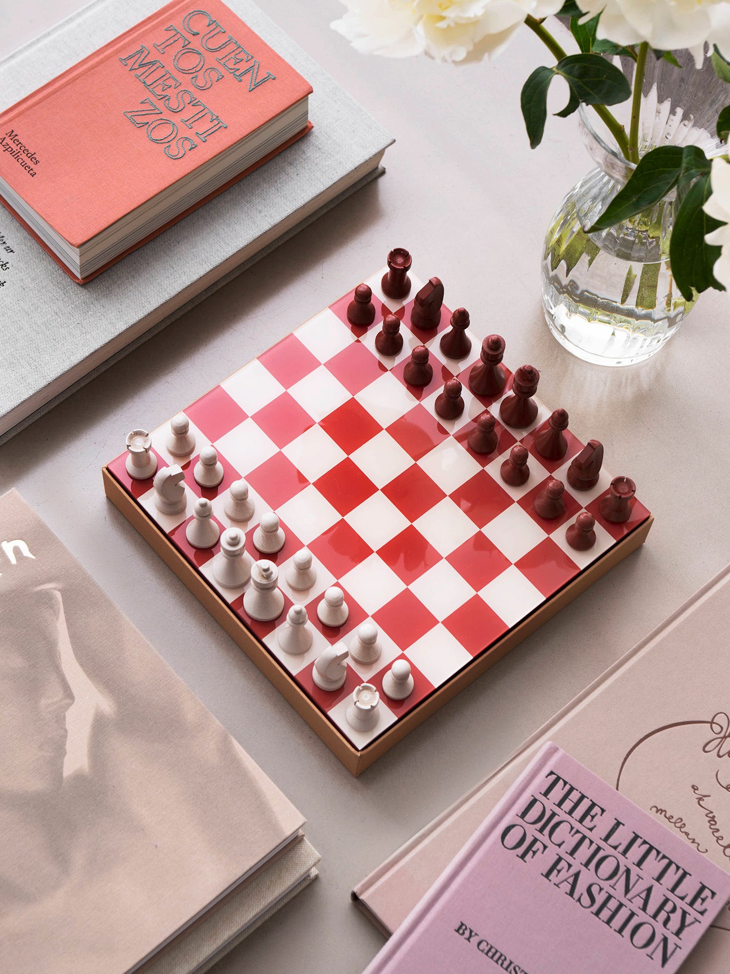 A red and white checkered chessboard with matching chess pieces arranged on starting position, waiting for the game to start, on a tabletop, surrounded by books and a glass vase with white flowers.