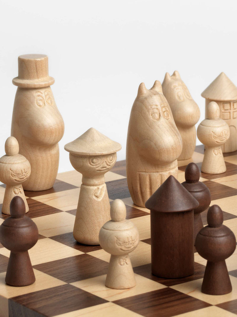 A close-up photo of a set of uniquely shaped wooden chess pieces in light untreated wood and dark untreated wood, arranged on a checkered wooden chessboard against a clean white background. The pieces depict Snufkin,  Moominpappa with his tall hat as the king and Moominmamma in striped apron as the queen, with Moomintroll and the round tall Moominhouse, with several Little My figures, playing as the white pieces against the dark Moomin house as the tower and three Little Mys as pawns.