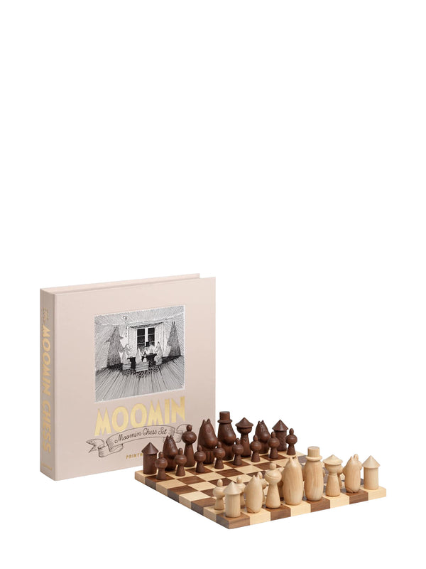 A wooden chess set with two-tone light and dark brown pieces arranged on a checkered board ready for play, next to a beige box titled "MOOMIN" with gold and smaller title "Moomin Chess Set" on an illustrated banner, with a large illustration from the Moomin books or comics by Tove and Lars Jansson in black-and-white on the cover, photographed on a white background.