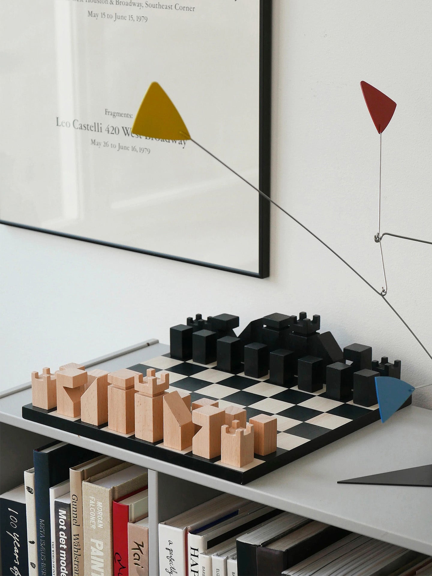 A black and white checkered chessboard with beige untreated wooden chess pieces arranged next to the black painted chess pieces, sitting on a bookshelf, alongside a colorful abstract metal sculpture and a framed print on a white wall.