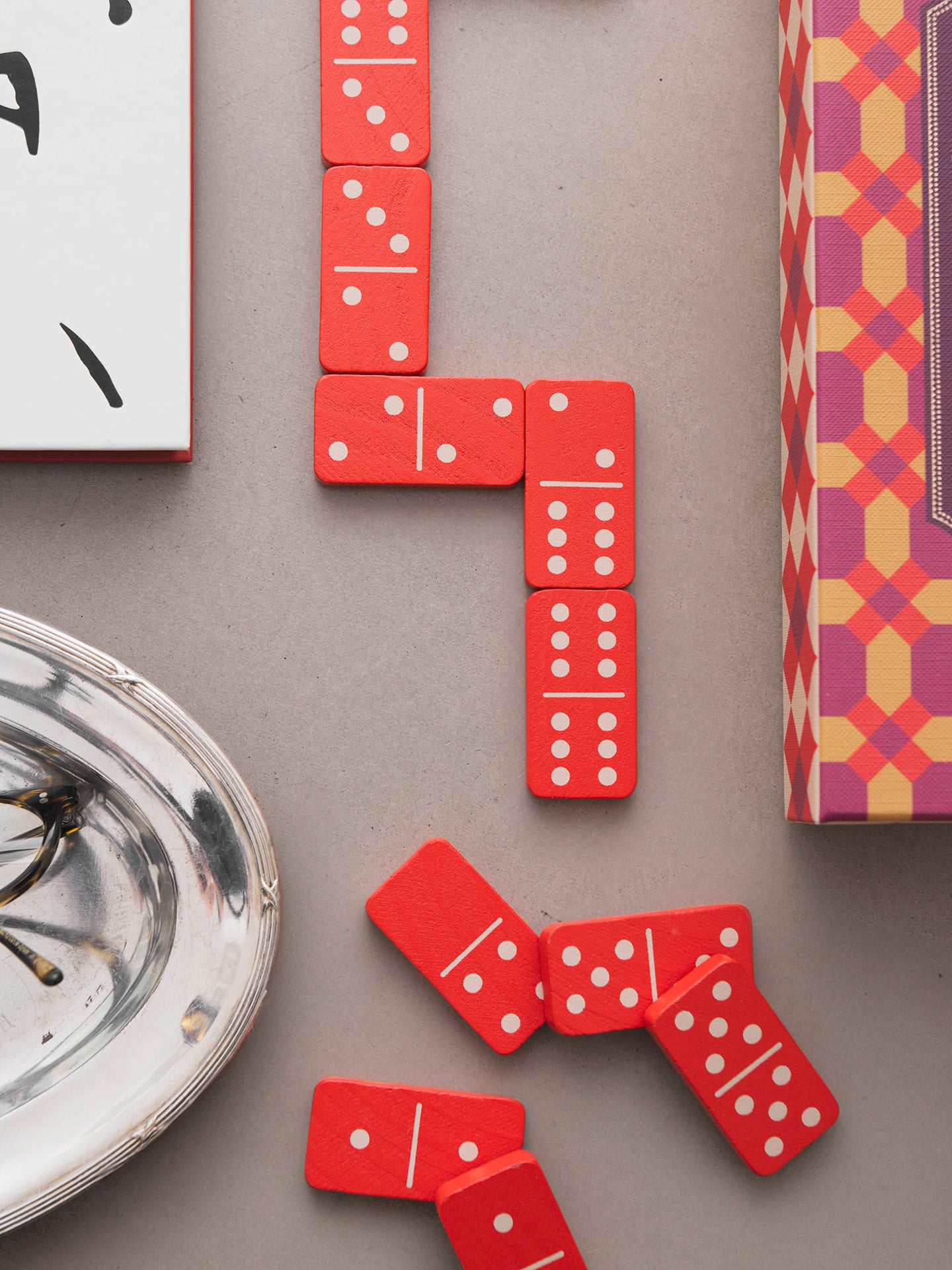 Red domino tiles with white pips arranged on a neutral gray surface, alongside partial views of patterned and white boxes and a silver dish.