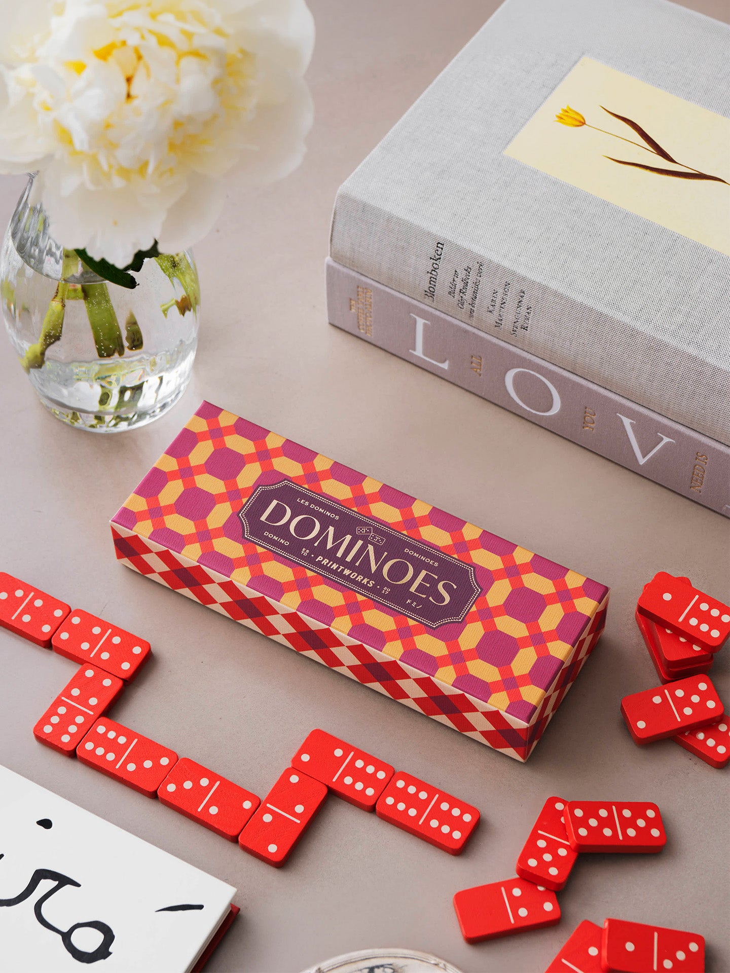 Dominoes game set with a patterned red and yellow box, red domino tiles, and a white peony in a glass vase on a beige tabletop.