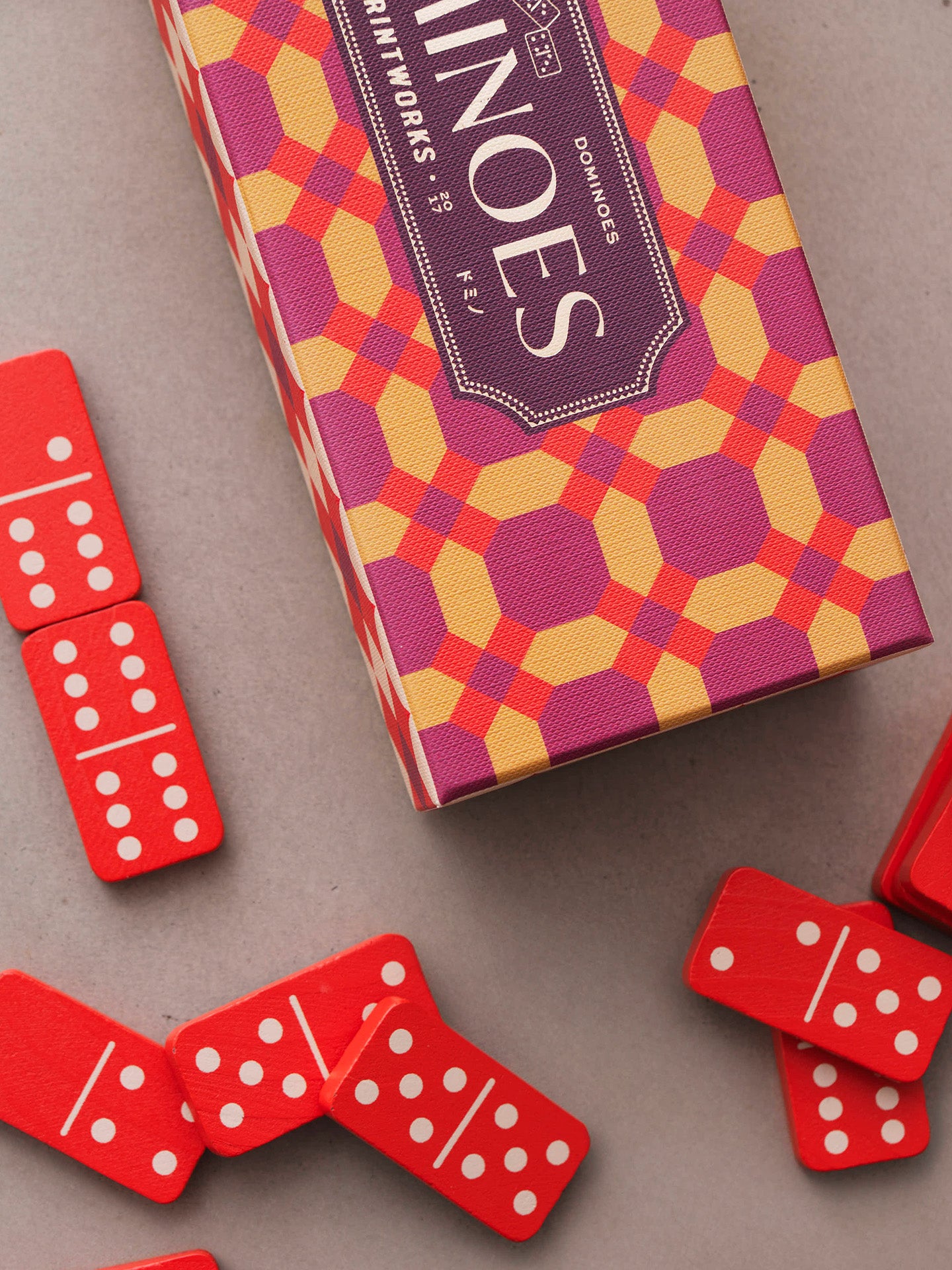 A colorful game box labeled 'Dominoes' with a geometric pattern, shown on a neutral gray surface with several red domino tiles scattered around it.