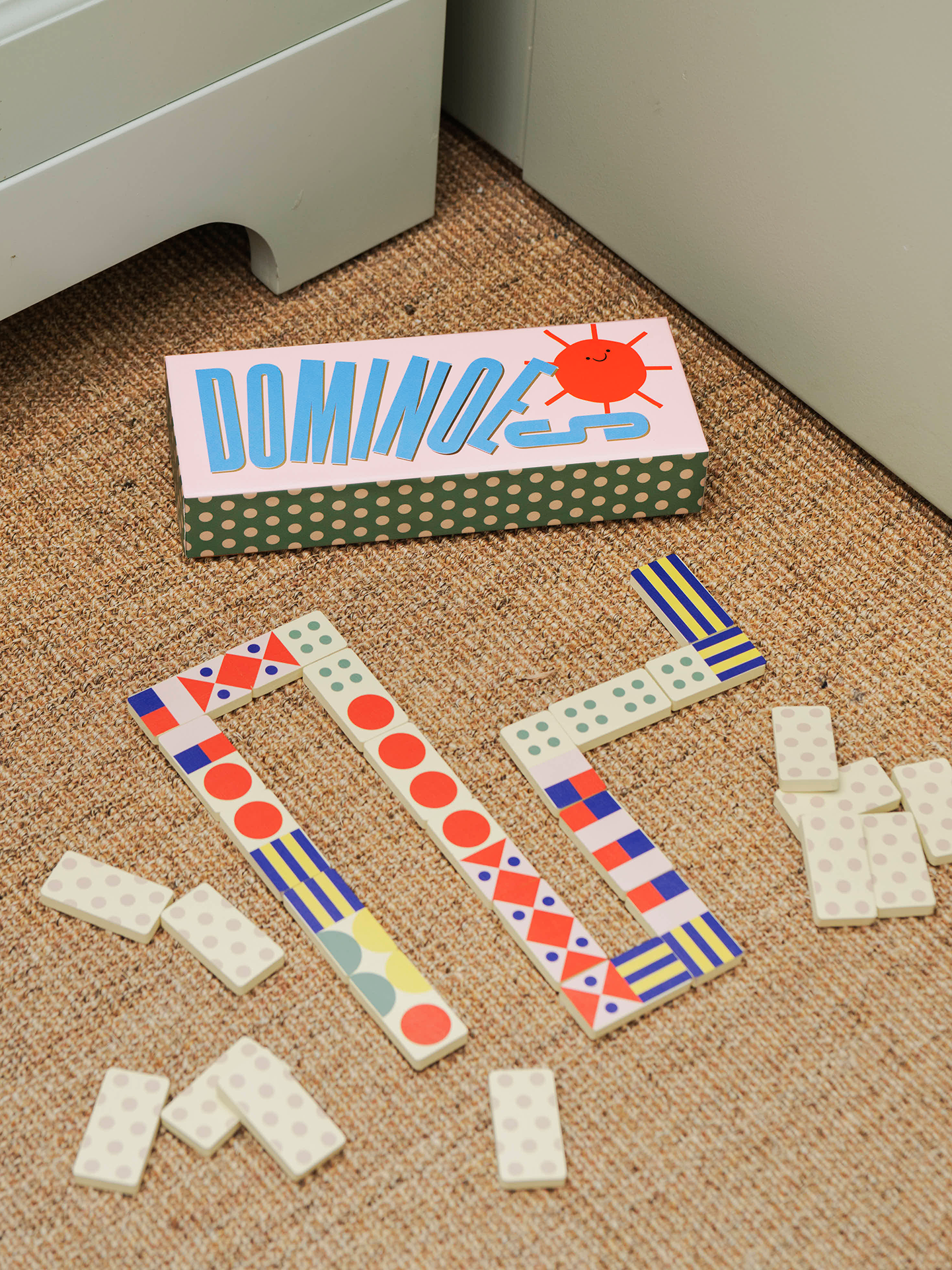 A set of colorful patterned domino tiles and a matching box on a woven jute rug near a white cabinet. The box has a pink top with blue 'DOMINOES' text and a red sun illustration, and the dominoes feature various geometric patterns in red, blue, green, yellow, and white.