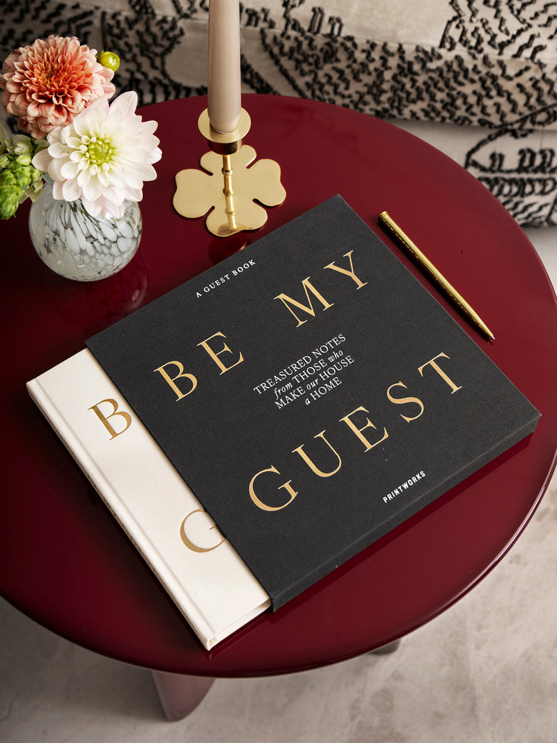 Ivory white notebook peaking from its' dark slipcover with golden coloured headline 'Be My Guest' and subheadline 'Treasured notest from those who make our house a home' create a beautiful still life on a red table with flower vase, candle holder and a pen