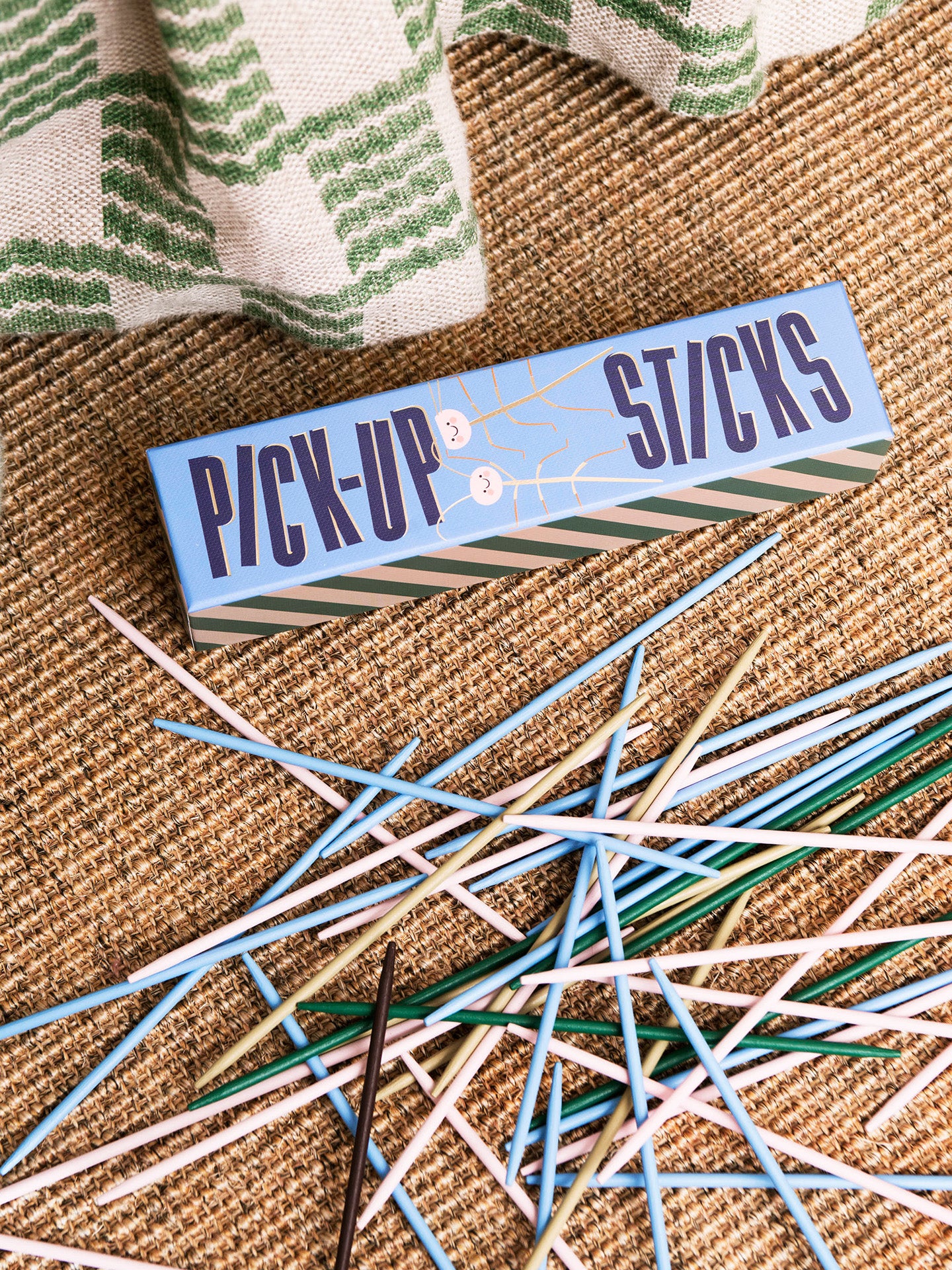 A light blue rectangular box labeled 'PICK-UP STICKS' sits on a woven tan surface with many loose wooden and plastic pick-up sticks scattered around it. A green-and-white striped cloth is partially visible at the top.