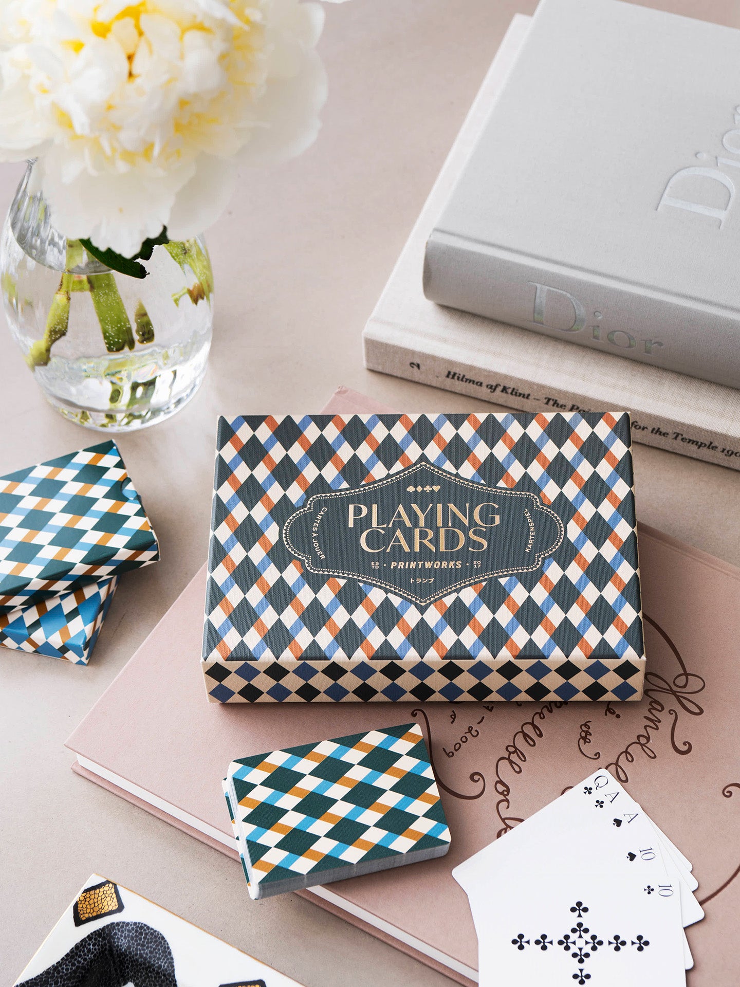 A boxed deck of patterned playing cards sits on a pink notebook with several cards fanned out, alongside matching folded card stacks and a glass vase with white flowers. The box reads 'PLAYING CARDS' and shows a geometric multicolor diamond pattern.
