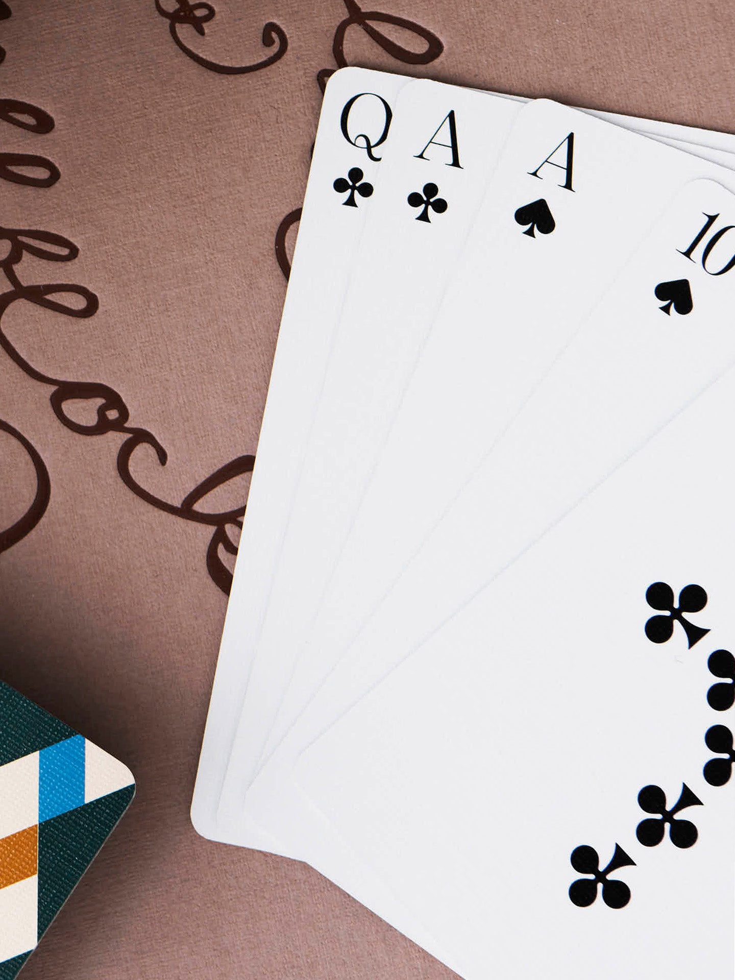 A fan of white playing cards showing the five cards from the black suits, starting from the left queen of clubs and the ace of clubs, and ace of spades and 10 of spades, with an unknown card from the clubs, laid on a brown textured surface with decorative script and a slice of diamond tile patterned card back at the lower corner.