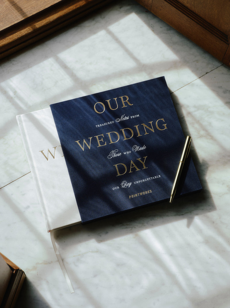 Off-white guest book in darker slipcover titled 'Our Wedding Day' written on gold letters placed on a marble window sill with a silver pen