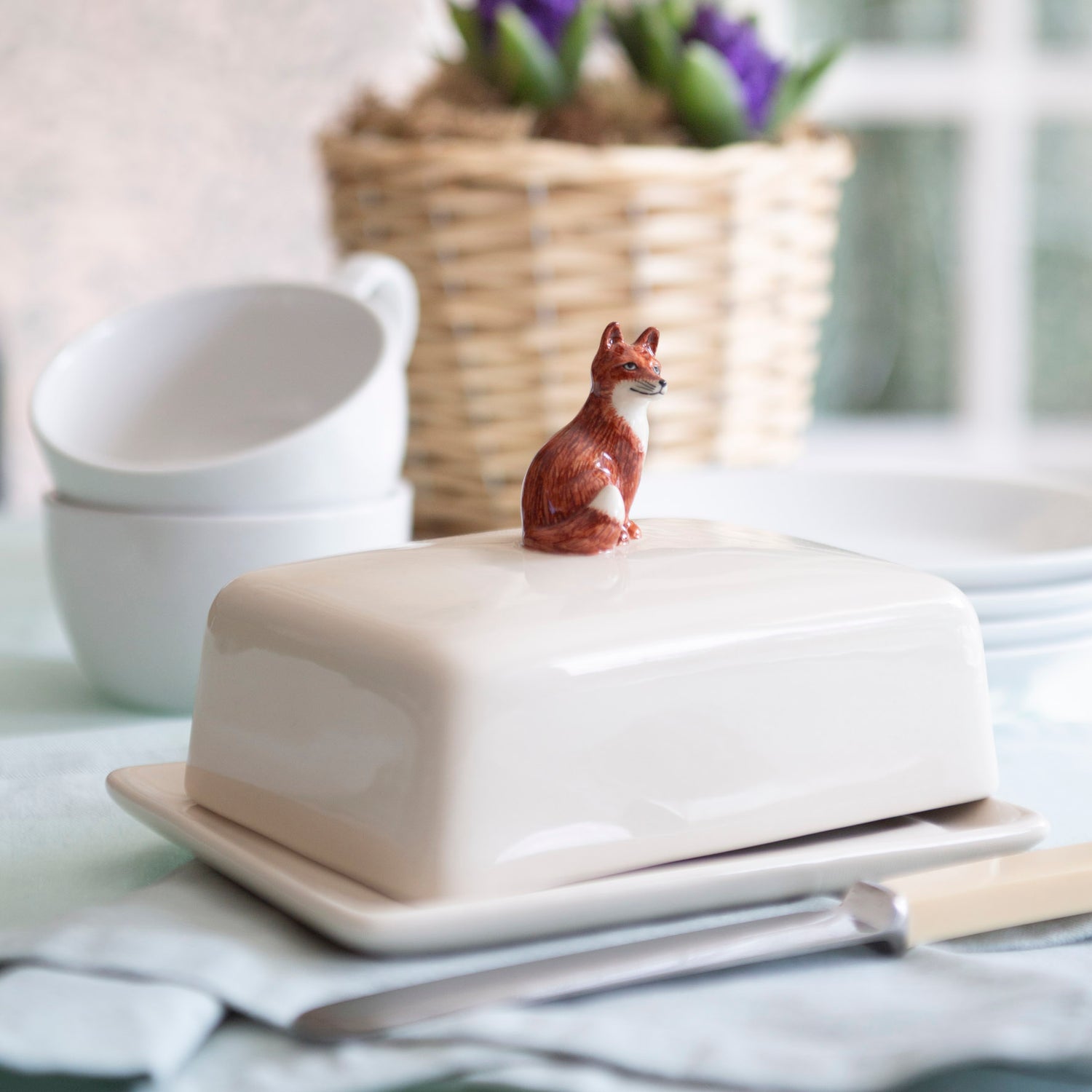 Butter dish with a fox figurine on top, set on a kitchen counter with bowls and a basket in the background.