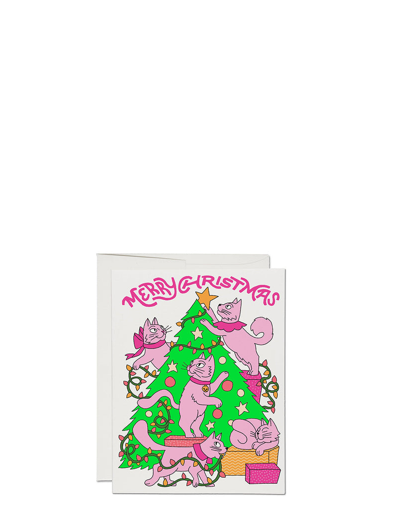 A Christmas greeting card featuring pink cats decorating a green Christmas tree with presents and lights, with the text 'MERRY CHRISTMAS' in pink above. The card is shown with a white envelope behind it on a plain white background.