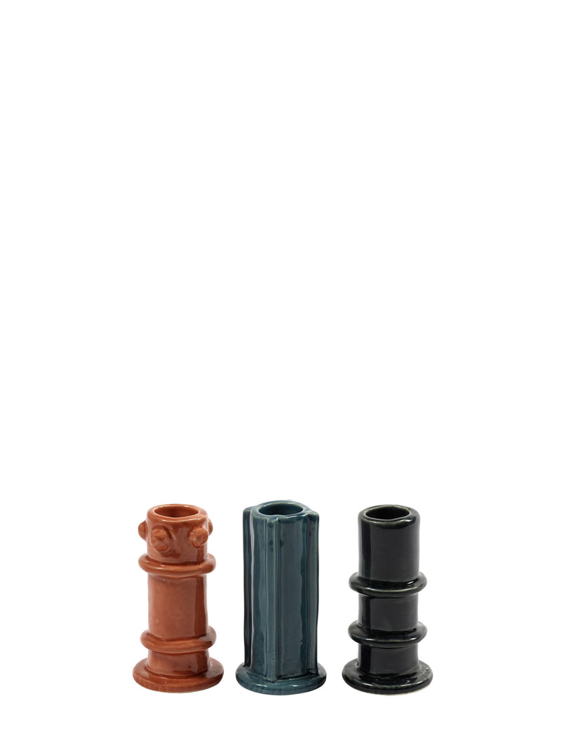 Three slightly different glazed ceramic candle holders in rust orange, teal green, and charcoal black from Serax on a white background