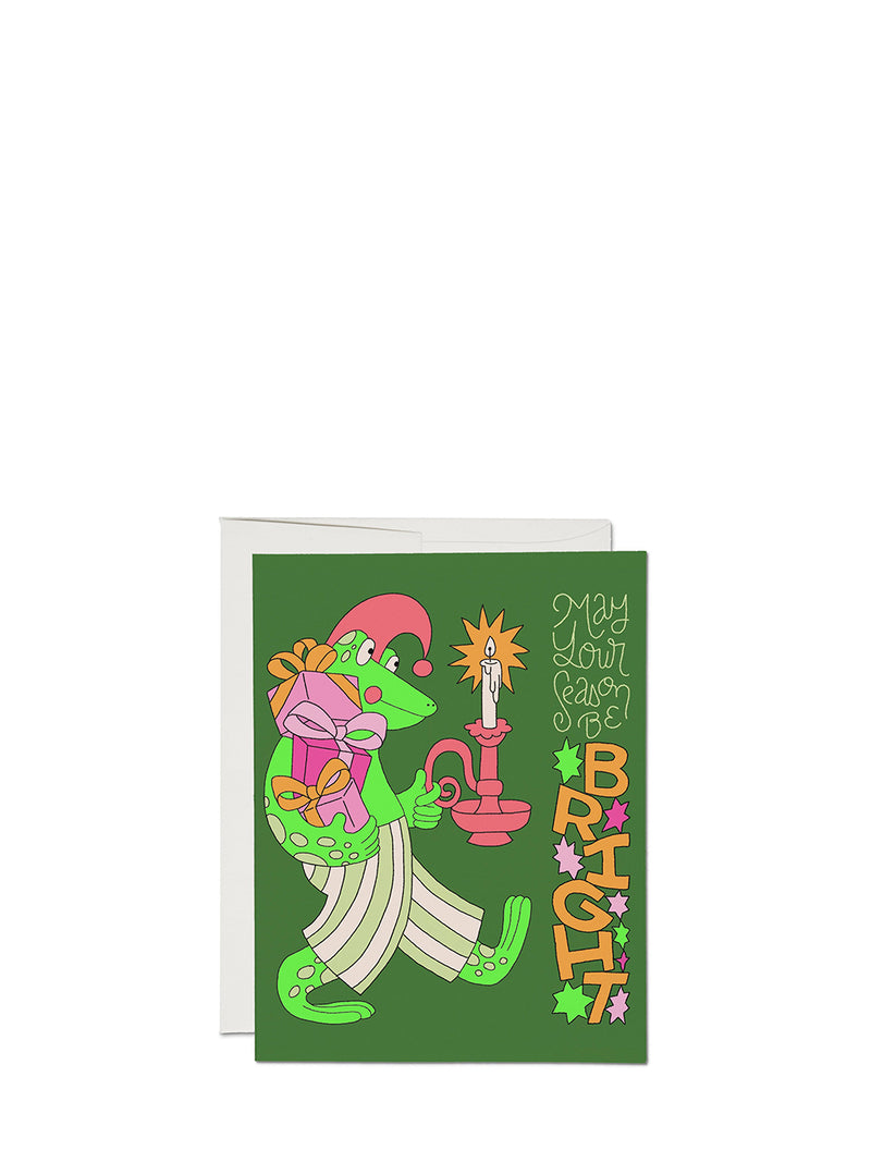 A green greeting card with a whimsical cartoon frog dressed in pink and white striped pajamas, holding a pink candle, next to a white envelope on a white background.