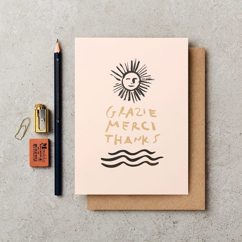 Greeting card with 'Grazie Merci Thanks' text and sun design on a concrete surface with stationery items.