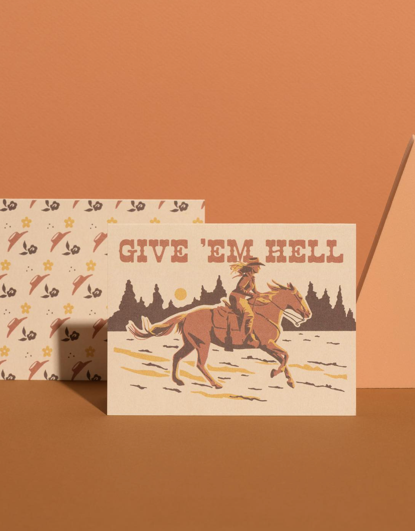 Two greeting cards on a brown background, one with a cowboy design and 'Give 'em Hell' text, the other with a pattern of arrows and stars.