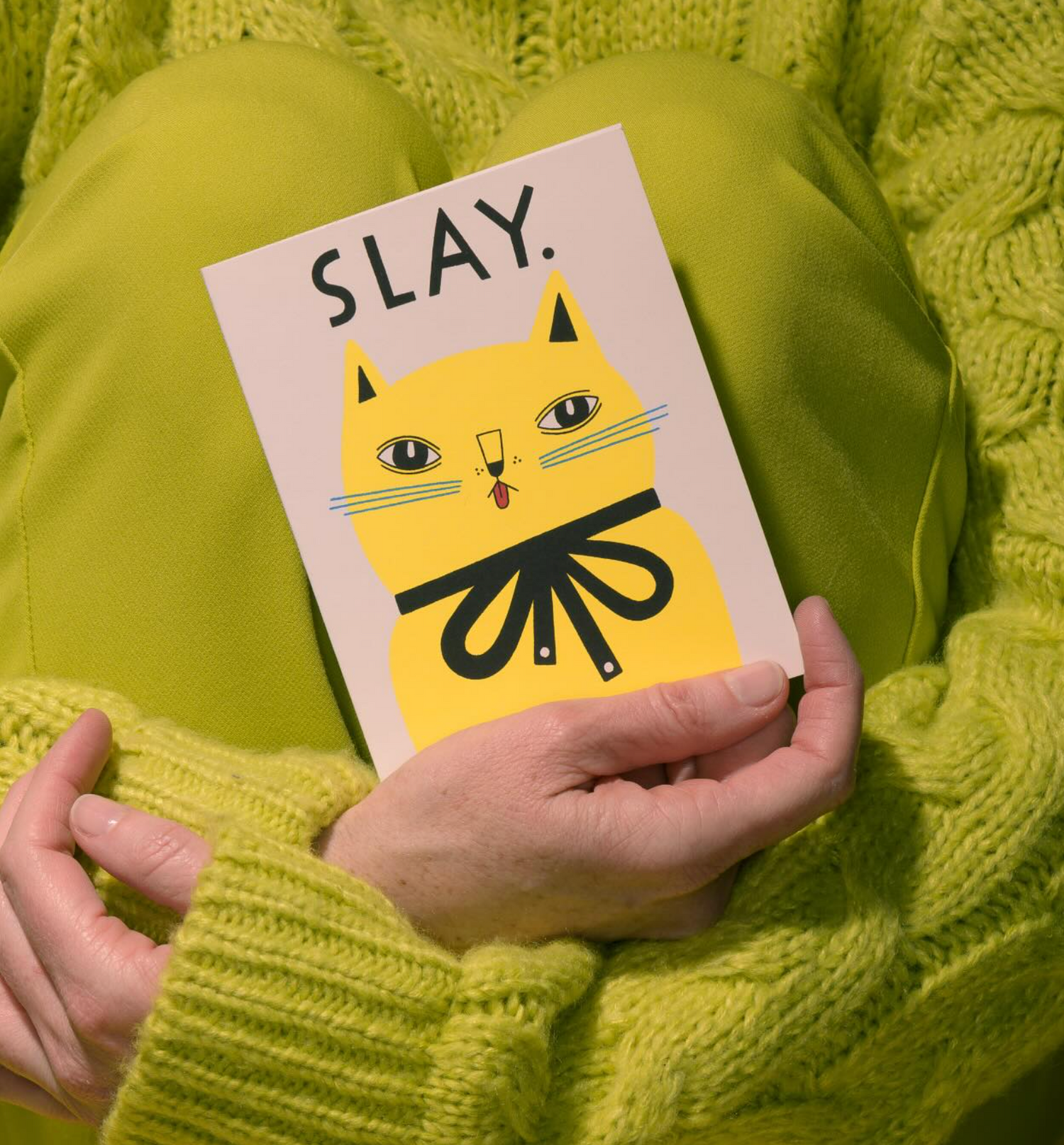 Person holding a card with a cat illustration and 'SLAY' text, wearing a green sweater.