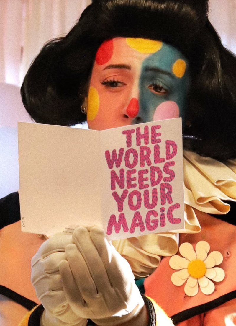 Person with colorful makeup holding a sign that says 'The World Needs Your Magic' with a white background.