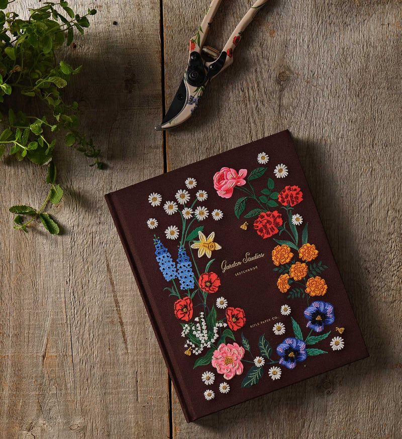 Notebook with floral embroidery on a wooden surface with gardening tools and plants.