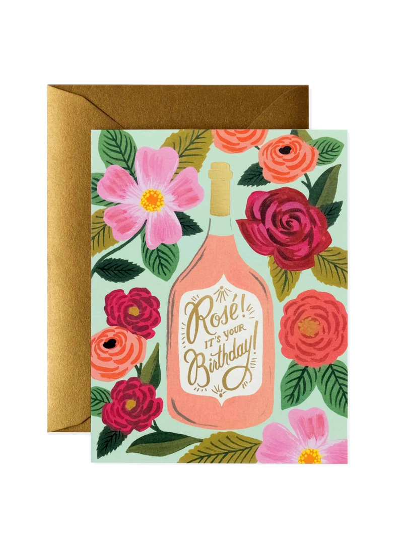 A birthday card with a floral pattern and a bottle illustration, with a envelope behind it.