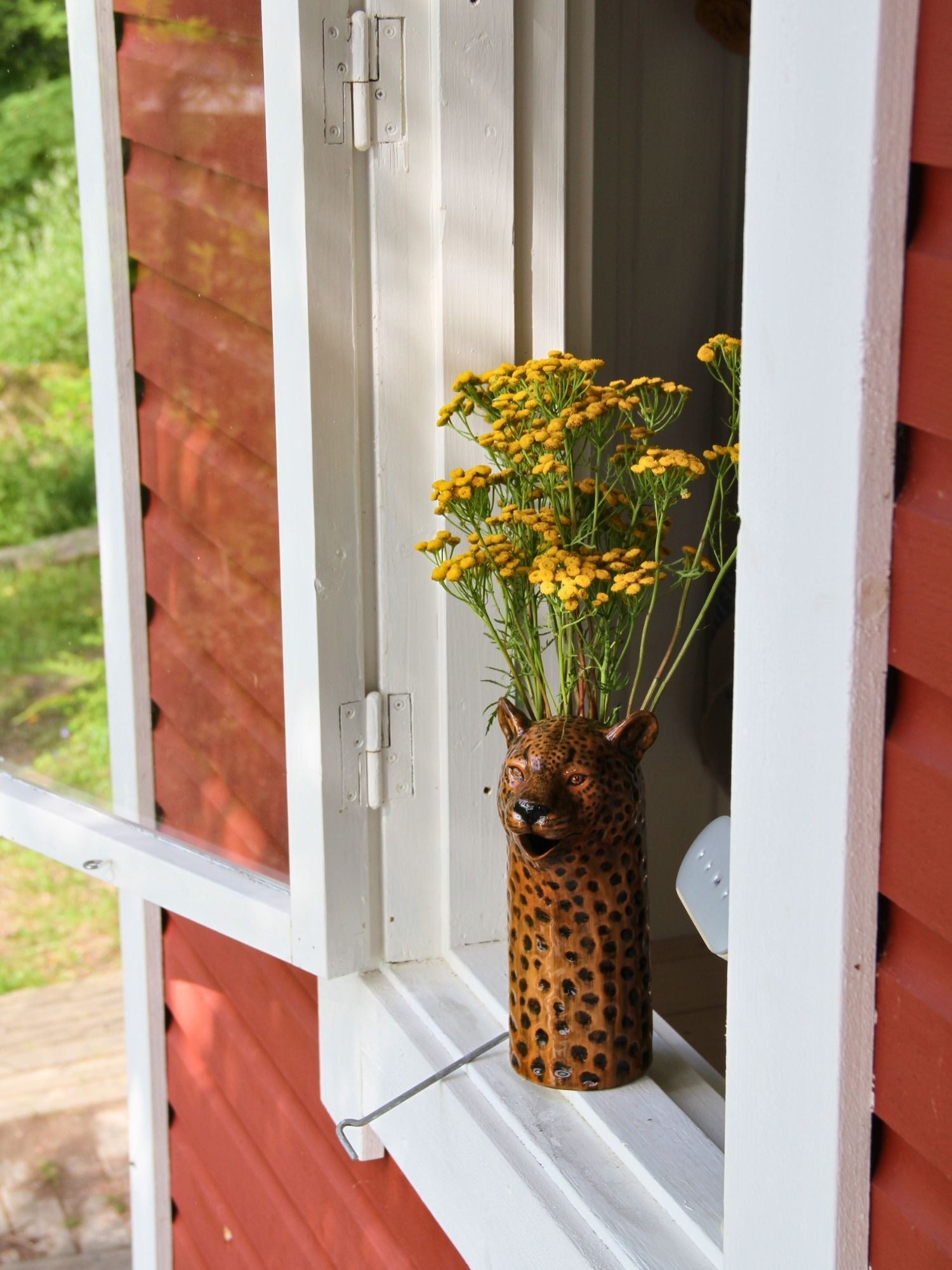 A decorative ceramic vase shaped like a leopard head with a spotted orange-and-black pattern, holding yellow flowers, set on a white window sill against a red wooden wall.