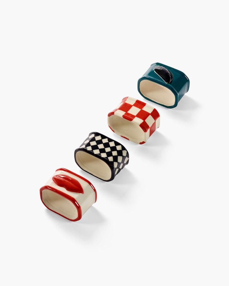 Napkin rings in line, one dark teal with a black mussel relief emblem, red and off-white checkered, third one with black and off-white diagonal checkerboard pattern and fourth with red edges and red lips relief.