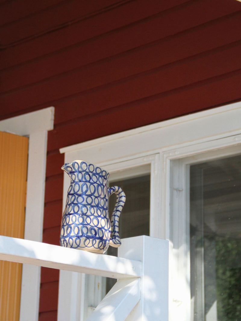 A blue and white patterned ceramic pitcher resting on a white wooden railing in front of a red house exterior with white trim.